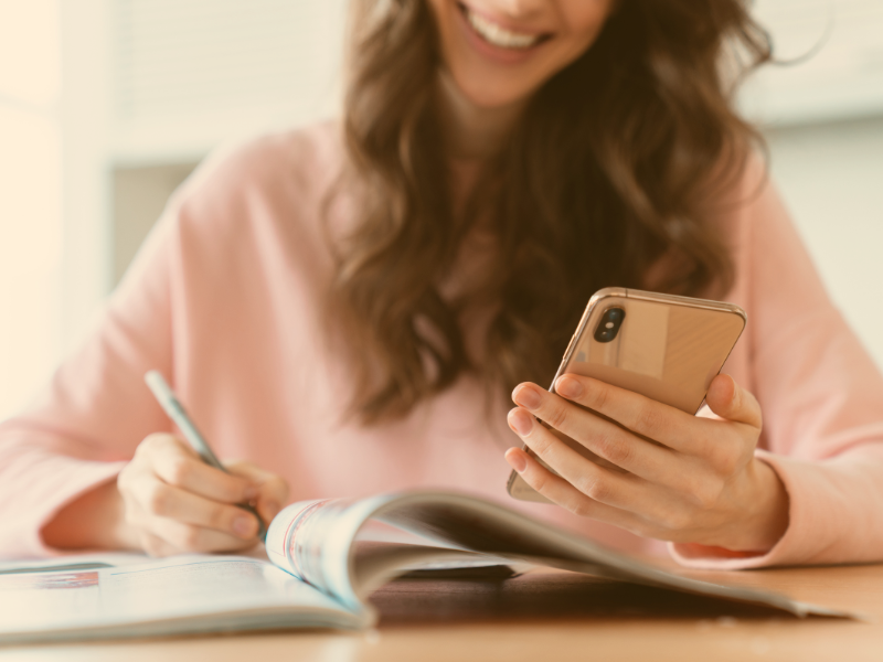 a woman in a pink sweater with brown hair looking at phone smiling and writing something in a notebook