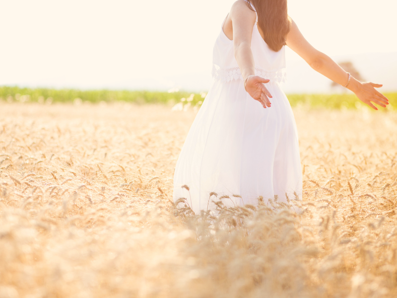 a woman in a white dress walking though a field of wheat in the sunshine